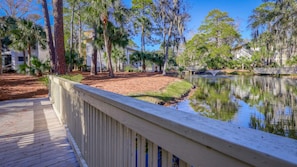 A wooden deck with a railing overlooking a calm water body, surrounded by trees and buildings. - MyStay at 3057 Seascape | South Forest Beach | Private Beach Cabana | Sleeps 7 (Hilton Head Island)