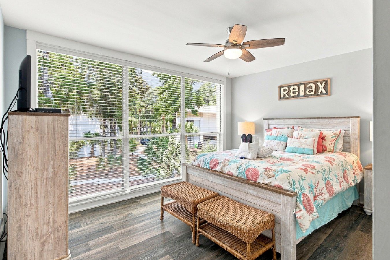 A bedroom with a bed, a ceiling fan, a large window with blinds, a wooden headboard, and a 'relax' sign on the wall.
