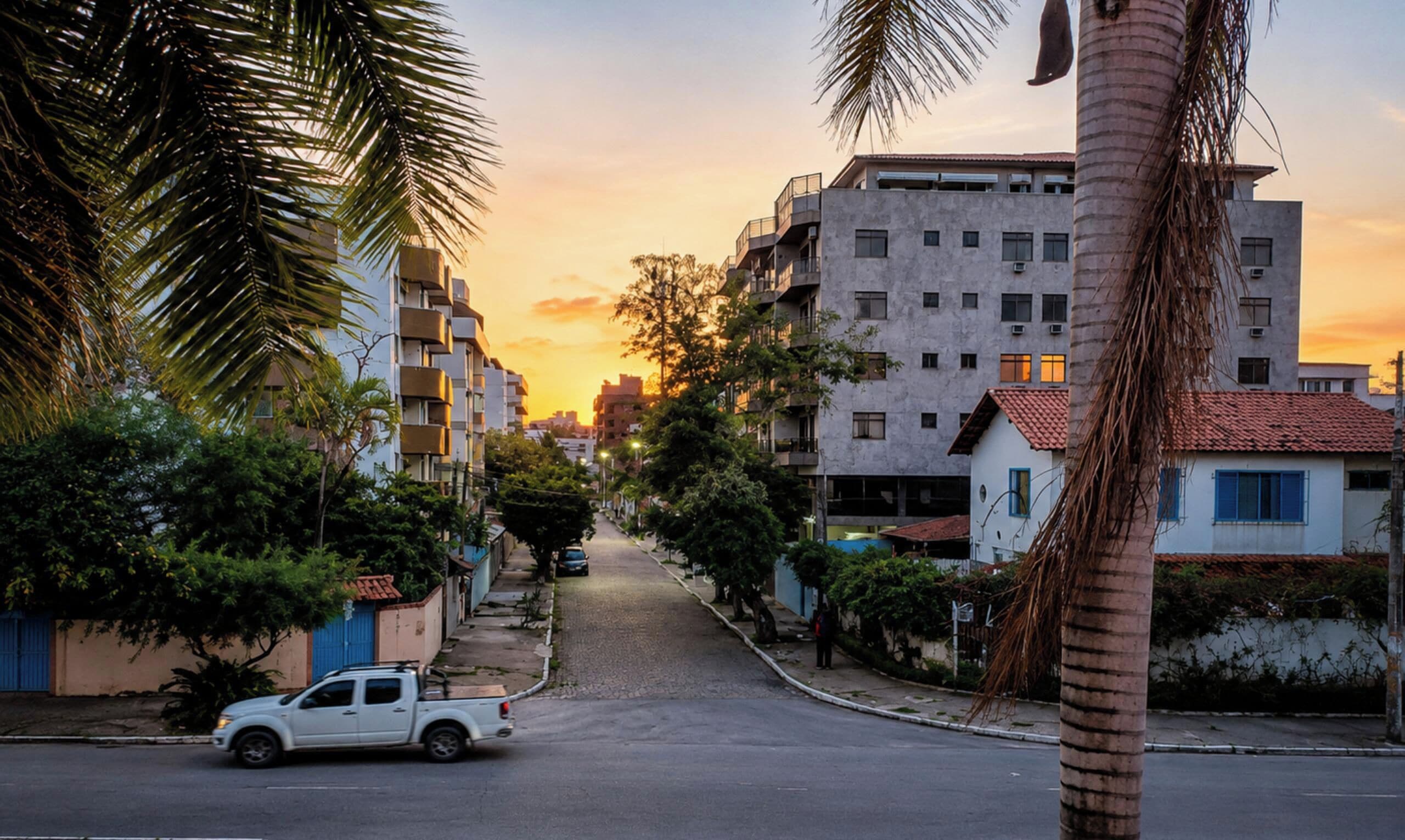 Vista de la calle con un auto blanco estacionado al lado, edificios con balcones y palmeras a lo largo de la vía.