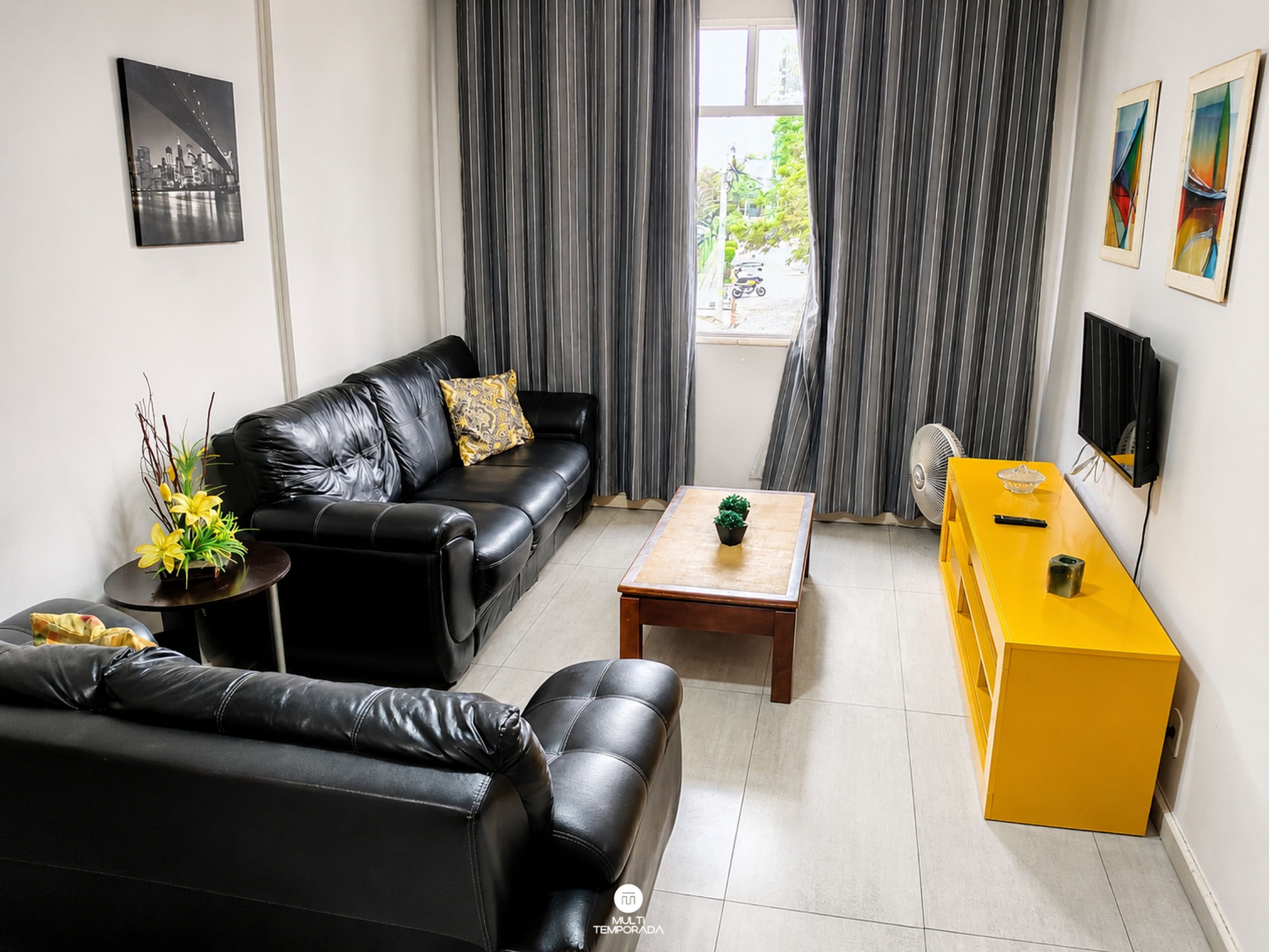 Un salon moderne avec un canapé en cuir noir, une table basse en bois, une petite table d’appoint jaune et un téléviseur fixé au mur.