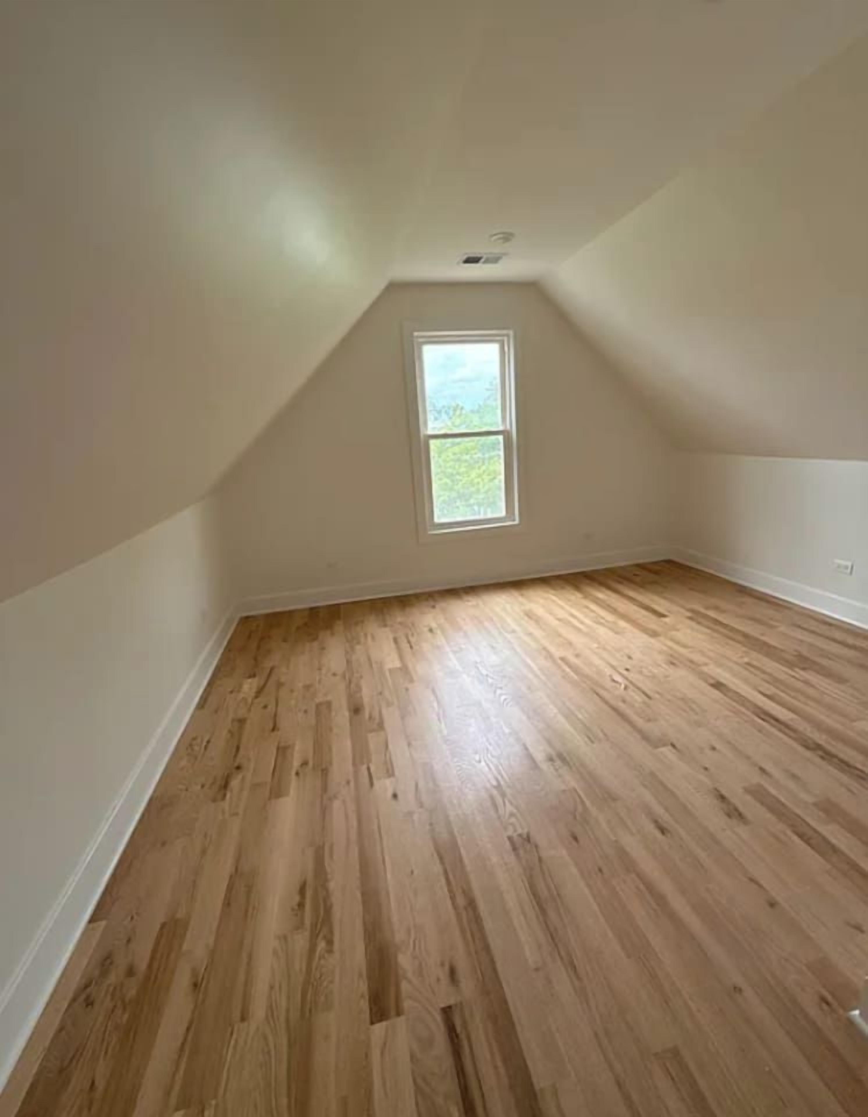 A room with wooden flooring, a sloped ceiling, and a window.