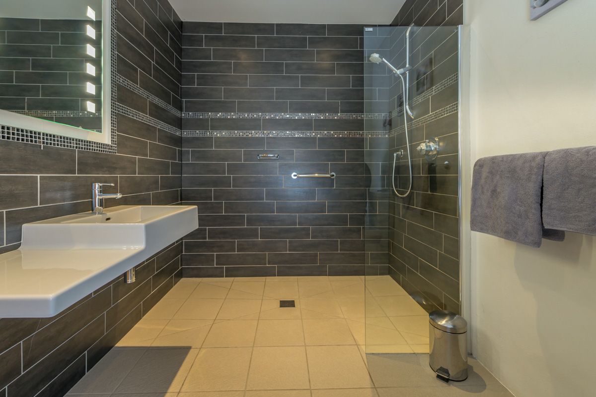 Un baño moderno con ducha de vidrio, lavamanos blanco y paredes de azulejos oscuros.