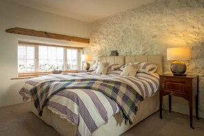 A bedroom with a large bed, two bedside tables with lamps, a stone wall, and a window with blinds. - Self Catering For Large Groups on The Llŷn Peninsula | Mynachdy-Sarn (Pwllheli)