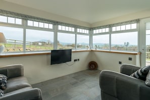 A room with a large bay window, a flat-screen TV mounted on the wall, and a stone-tiled floor. - Self Catering For Large Groups on The Llŷn Peninsula | Mynachdy-Sarn (Pwllheli)