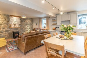A cozy living area with a stone fireplace, leather sofa, and a dining table set with a vase of flowers. - Holiday Cottage On A Working Farm On The Llŷn Peninsula | Ysgubor-Sarn (Pwllheli)