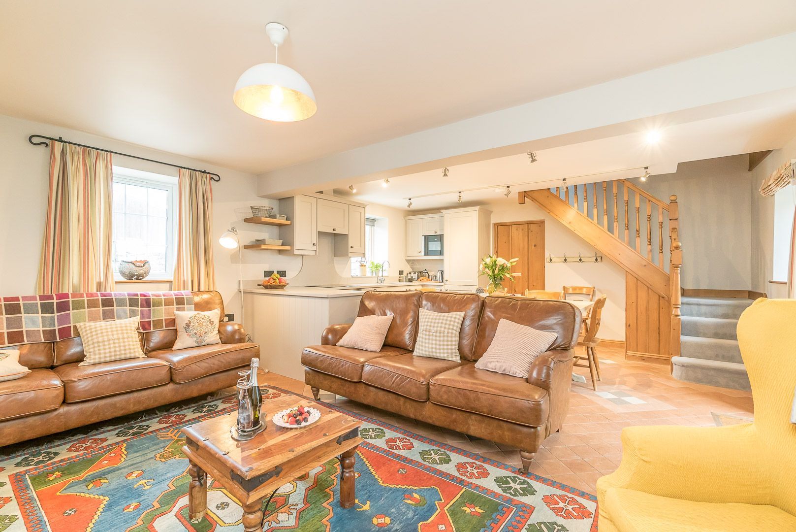A spacious living room with a sofa set, a coffee table, and a vibrant rug. A kitchen is visible in the background with a dining area and a staircase leading to another level.