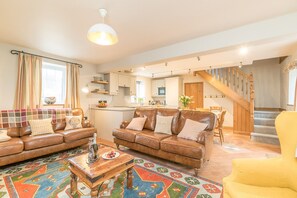 A spacious living room with a sofa set, a coffee table, and a vibrant rug. A kitchen is visible in the background with a dining area and a staircase leading to another level. - Holiday Cottage On A Working Farm On The Llŷn Peninsula | Ysgubor-Sarn (Pwllheli)