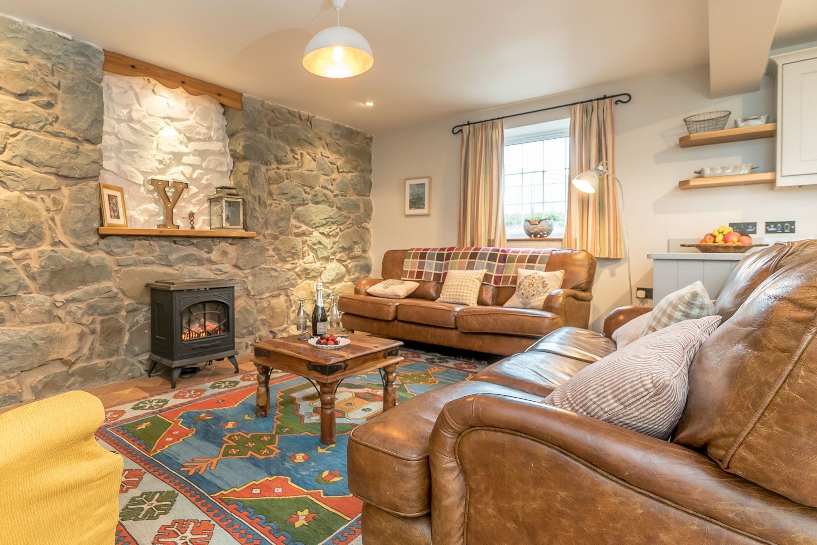 A cozy living room with a stone wall, a fireplace, a brown leather sofa, and a colorful rug.
