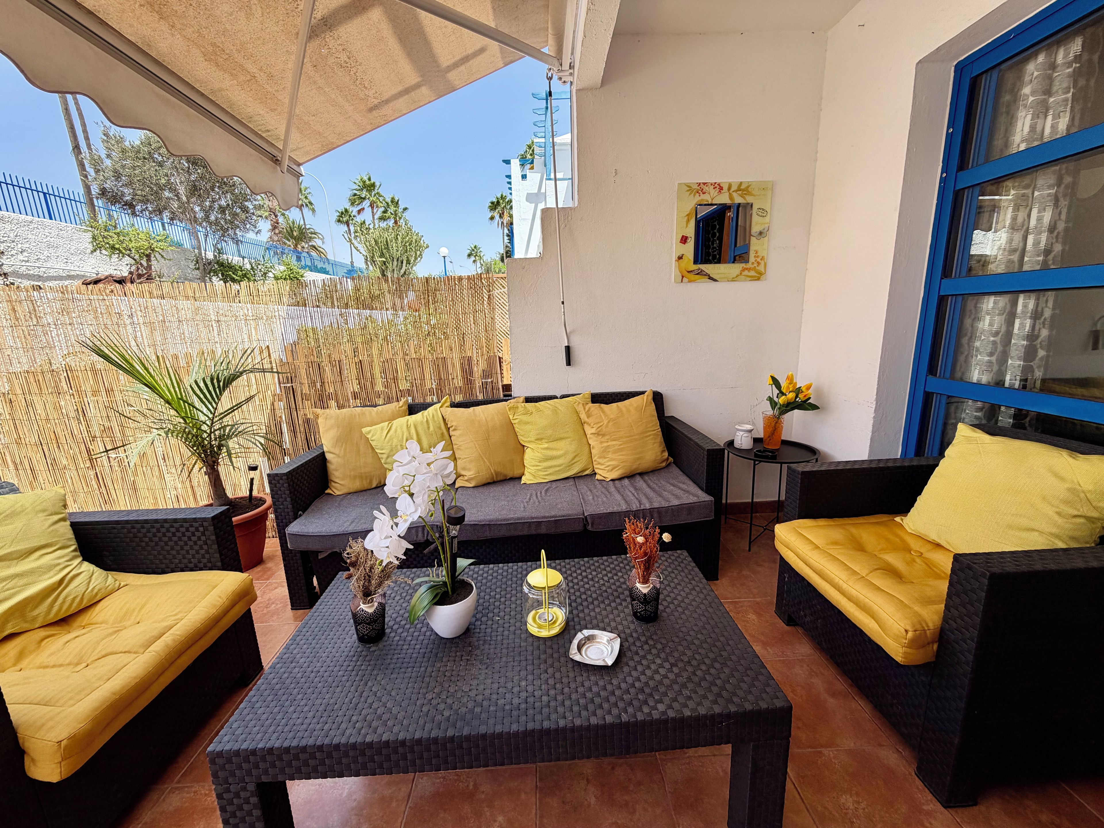 Eine sonnendurchflutete Terrasse mit einem grauen Sofa, gelben Kissen und einem Tisch mit Dekoration.