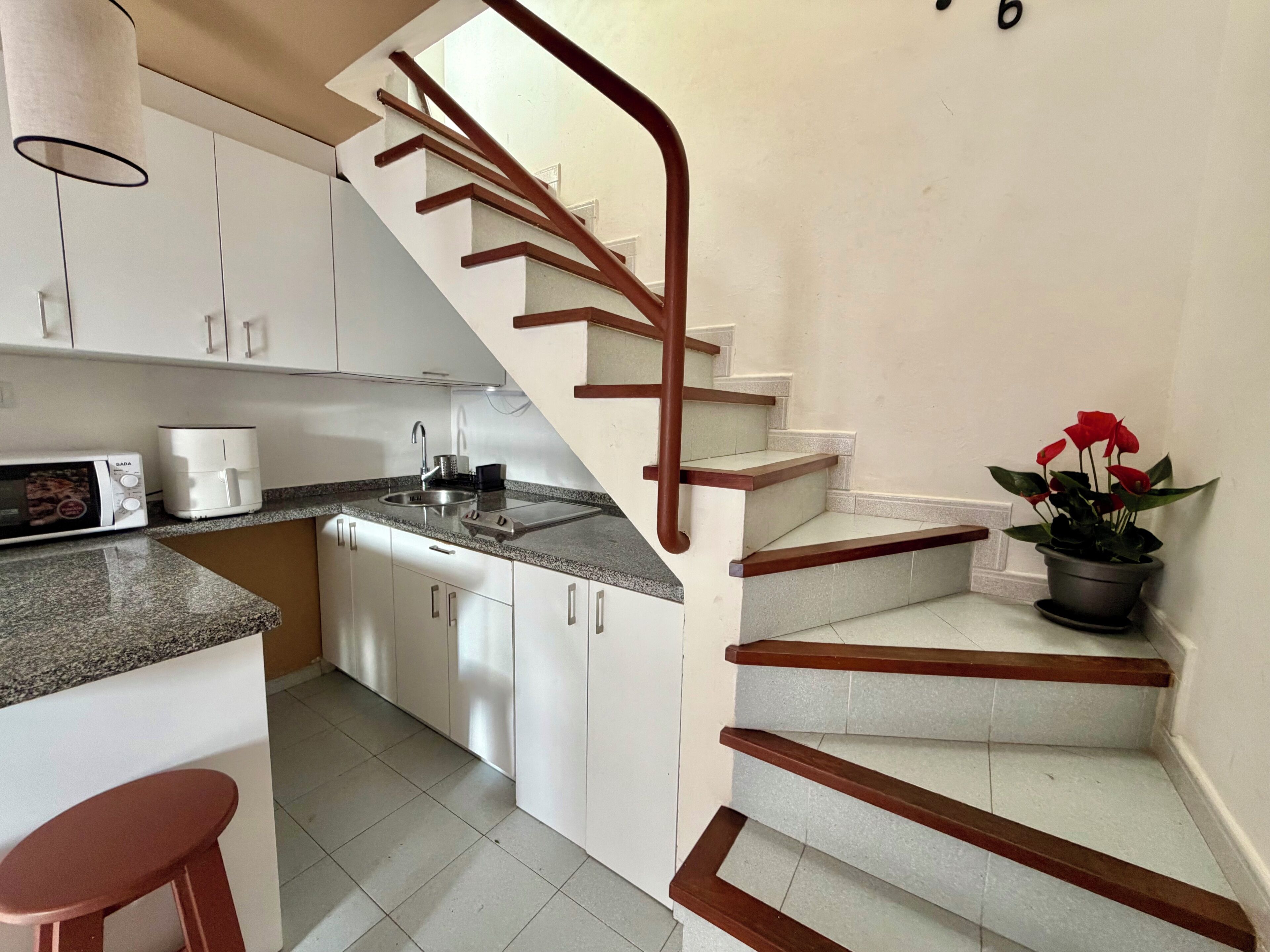 A kitchen with white cabinets, a sink, a microwave, and a stove. There is a wooden staircase with a handrail leading to an upper floor.