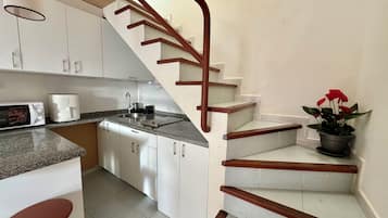 Une cuisine avec des meubles blancs, un évier, un four à micro-ondes et une cuisinière. On y trouve un escalier en bois avec une rampe menant à l’étage supérieur.