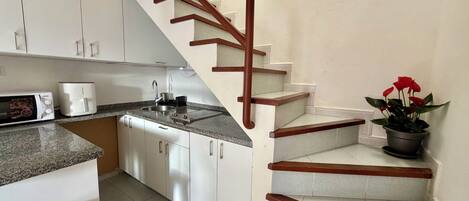 A kitchen with white cabinets, a sink, a microwave, and a stove. There is a wooden staircase with a handrail leading to an upper floor.