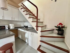 Une cuisine avec des meubles blancs, un évier, un four à micro-ondes et une cuisinière. On y trouve un escalier en bois avec une rampe menant à l’étage supérieur.