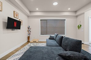 Ein modernes Wohnzimmer mit einem grauen Sofa, einem Flachbildfernseher, einem Fenster mit Jalousien und einem kleinen Tisch mit Blumen.
