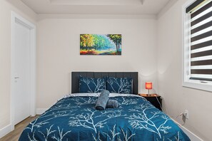 Ein Schlafzimmer mit einem Bett, einem Nachttisch, einer Lampe, einem Bild an der Wand und einem Fenster mit Jalousien.