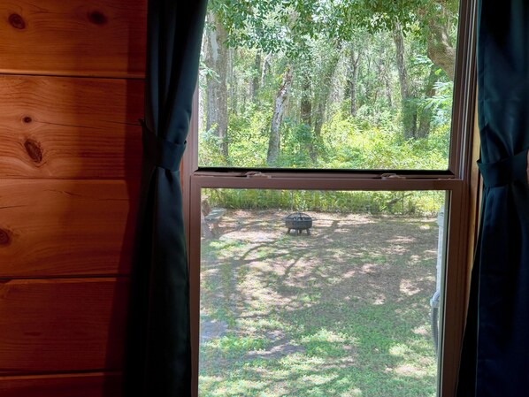 A window with a view of a forested area and a barbecue grill.