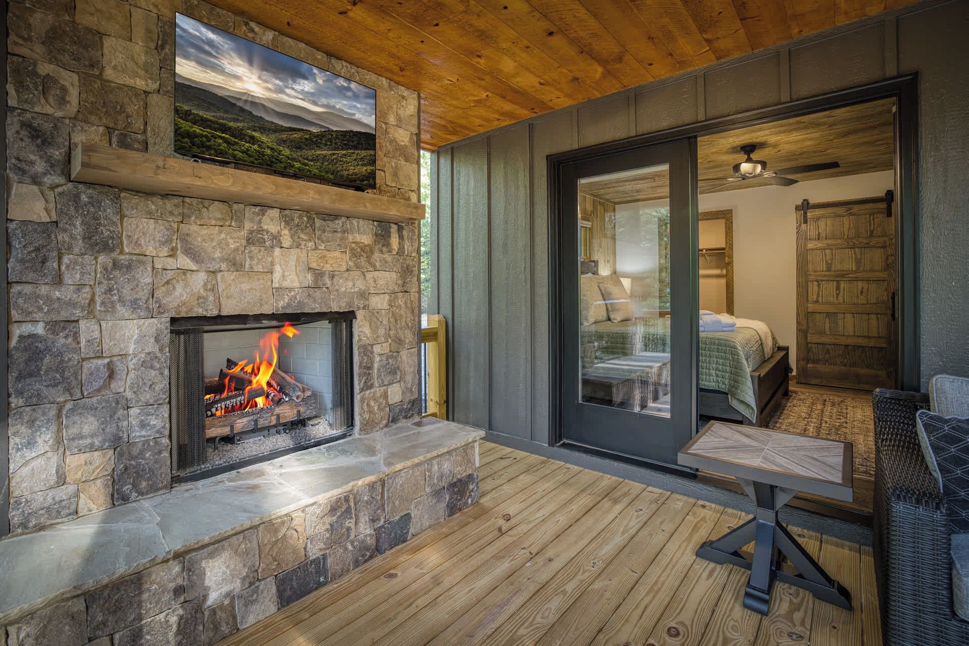 A stone fireplace with a fire, a wooden ceiling, and a view of a bedroom through a glass door.
