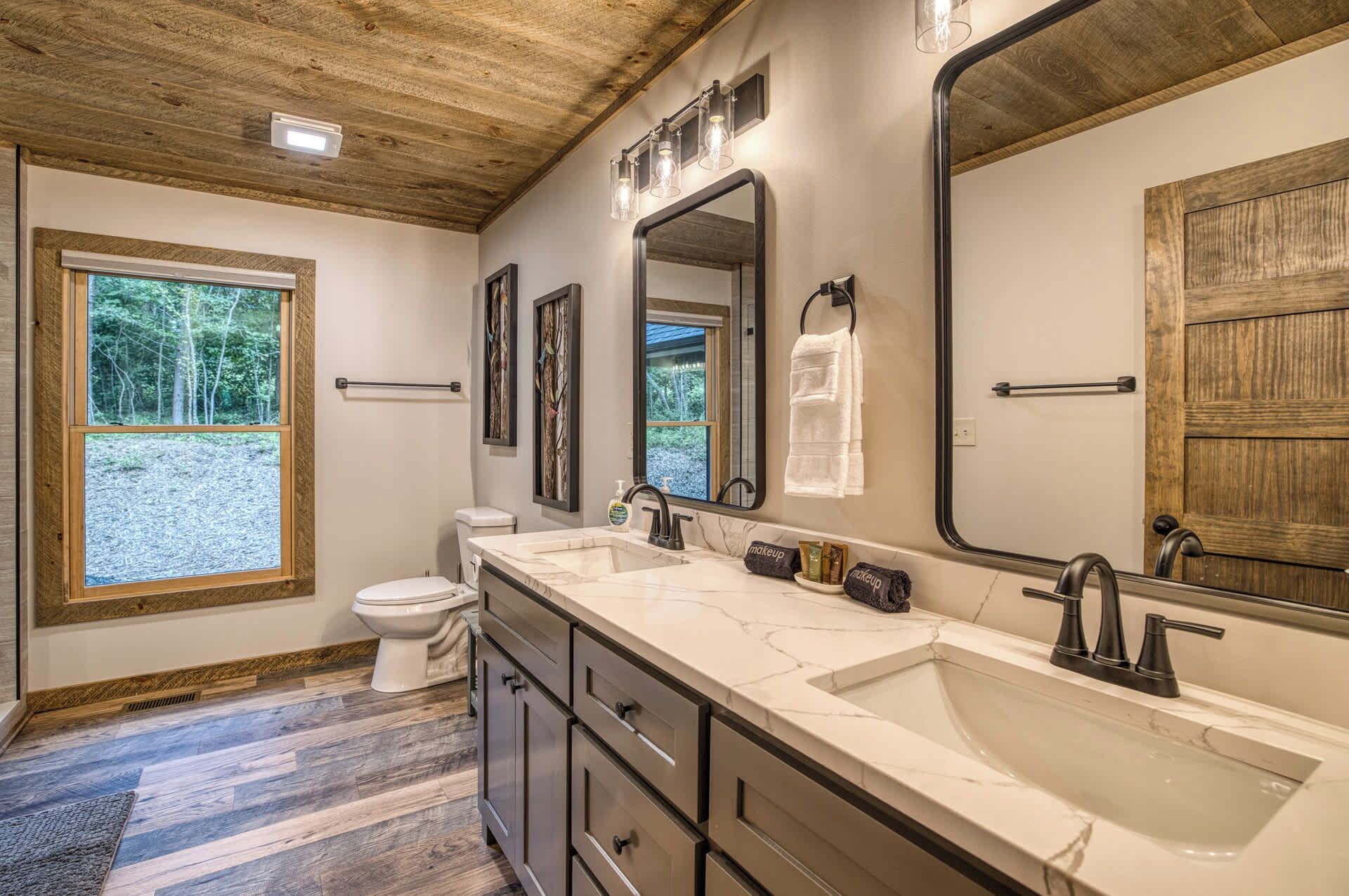 A bathroom with a large vanity, a double sink, a toilet, and a window with a view of trees.