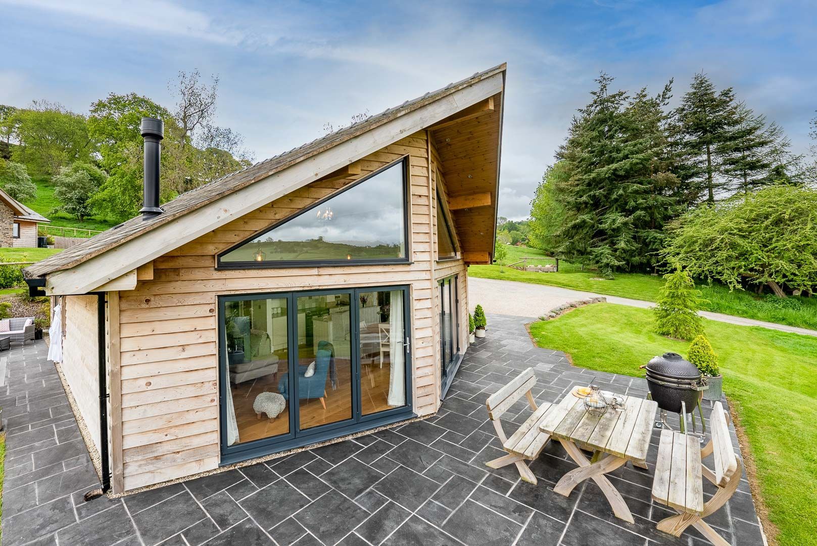 A modern wooden cabin with a sloped roof, large glass doors, and a patio with a table and chairs.