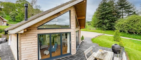 A modern wooden cabin with a sloped roof, large glass doors, and a patio with a table and chairs.