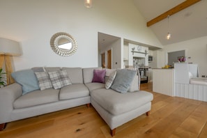 A modern living room with a sectional sofa, a decorative mirror, and a kitchen in the background. - Stylish Holiday Cottage In Rural Montgomery | Llety-Hiraeth (MONTGOMERY)