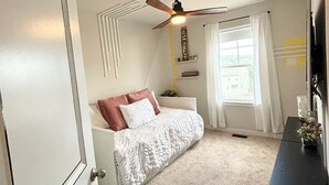 Une chambre à coucher avec un lit, un ventilateur de plafond, une fenêtre avec des rideaux et une commode.