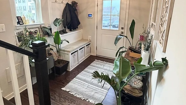Un couloir avec un escalier, un panneau d’accueil, un porte-manteau, une porte et divers pots de plantes.