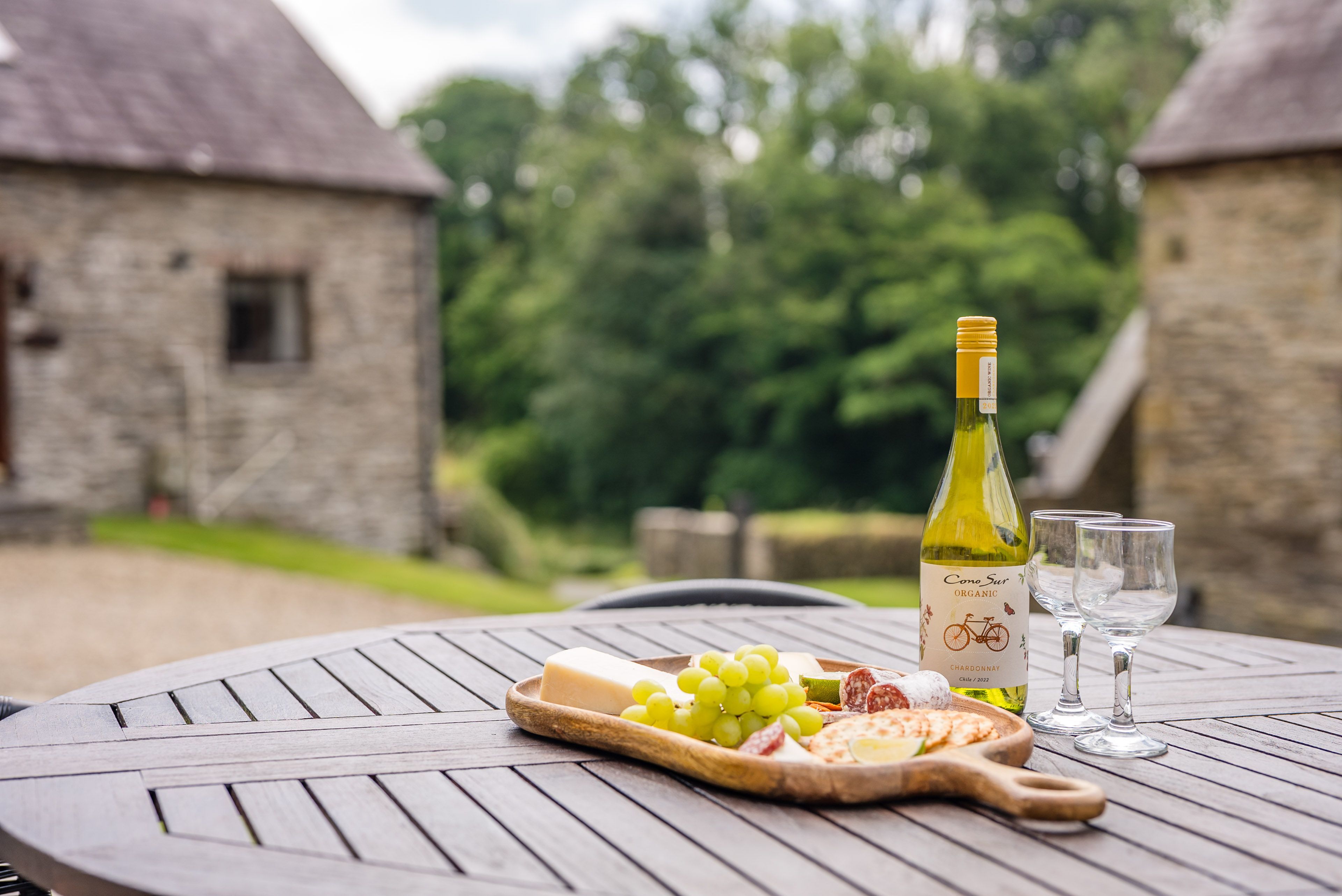 A table with a bottle of wine, glasses, a cheese board with grapes, and a wooden cutting board.