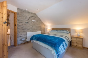 Ein Schlafzimmer mit schrägem Dach, einer Steinwand, einem Bett mit blauer Decke, zwei Nachttischlampen und einem Holznachttisch.