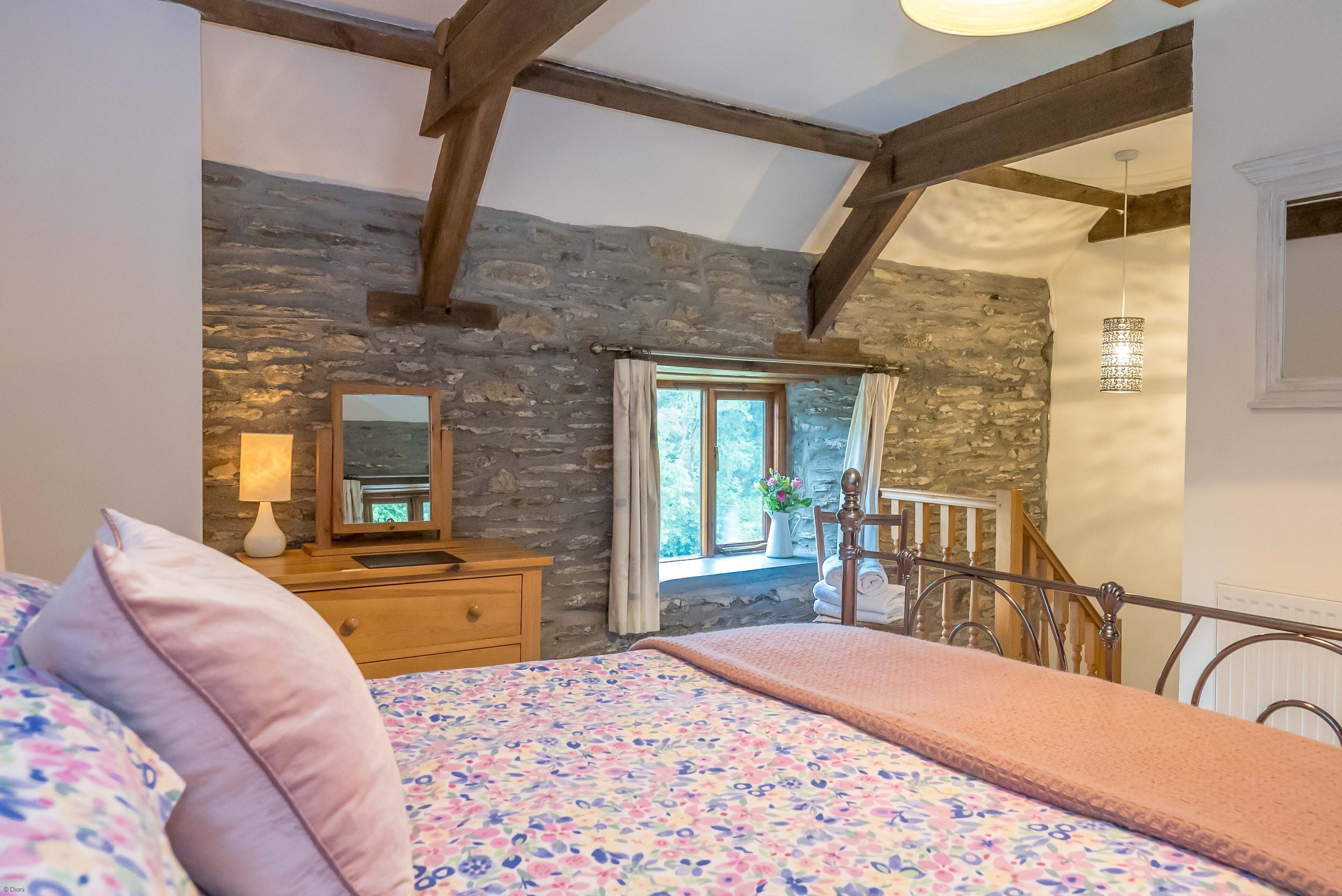 A bedroom with a stone wall, a wooden dresser, a bed with floral bedding, and a window with curtains.