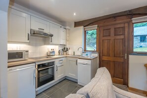 A compact kitchen with white cabinetry, a built-in oven, a dishwasher, and a window overlooking a garden. - One Bedroom Barn Conversion near Aberteifi | Certws-Y-Rhiw (Penparc)