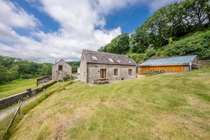 A stone house with a sloped roof and a wooden deck, surrounded by a grassy hill and trees. - A Converted Barn in a Beautiful Valley in Ceredigion | Granar-Y-Rhiw (Cardigan)