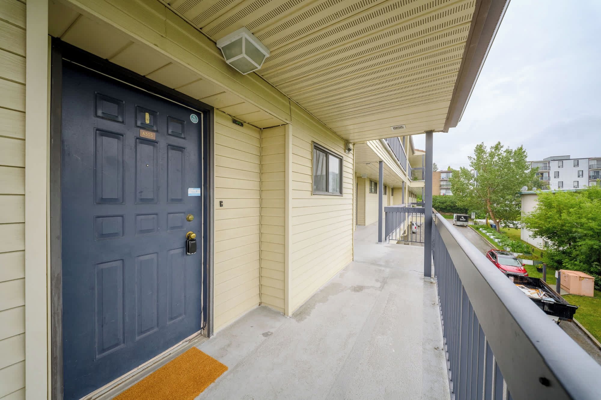 A residential building with a dark blue door, a covered balcony, and a view of a parking area with cars and greenery.