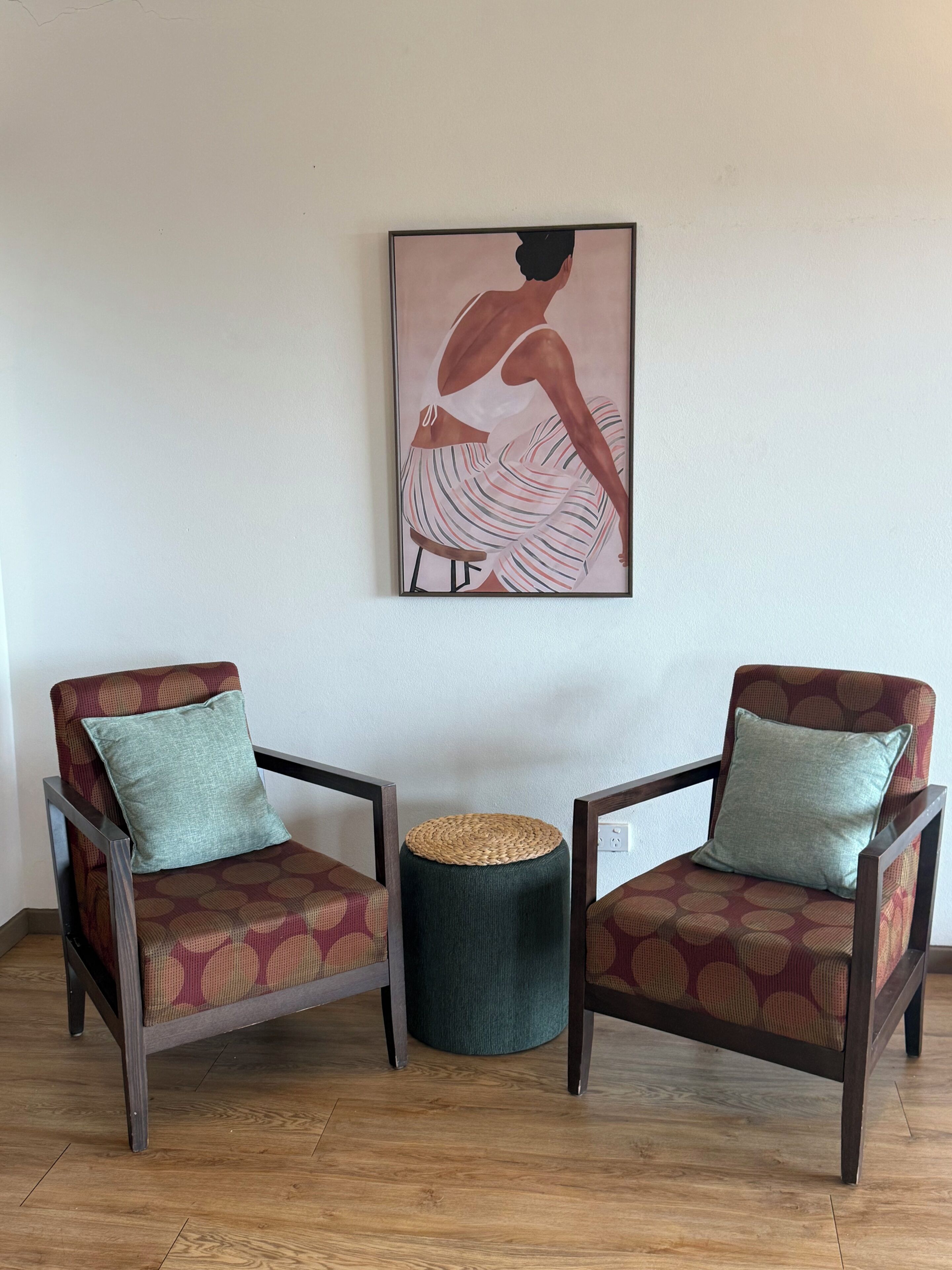 Deux fauteuils recouverts d’un tissu à motifs, un tabouret en osier et une œuvre d’art encadrée accrochée au mur.