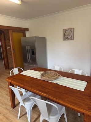 A dining area with a wooden table, white chairs, a woven basket, and a refrigerator in the background.