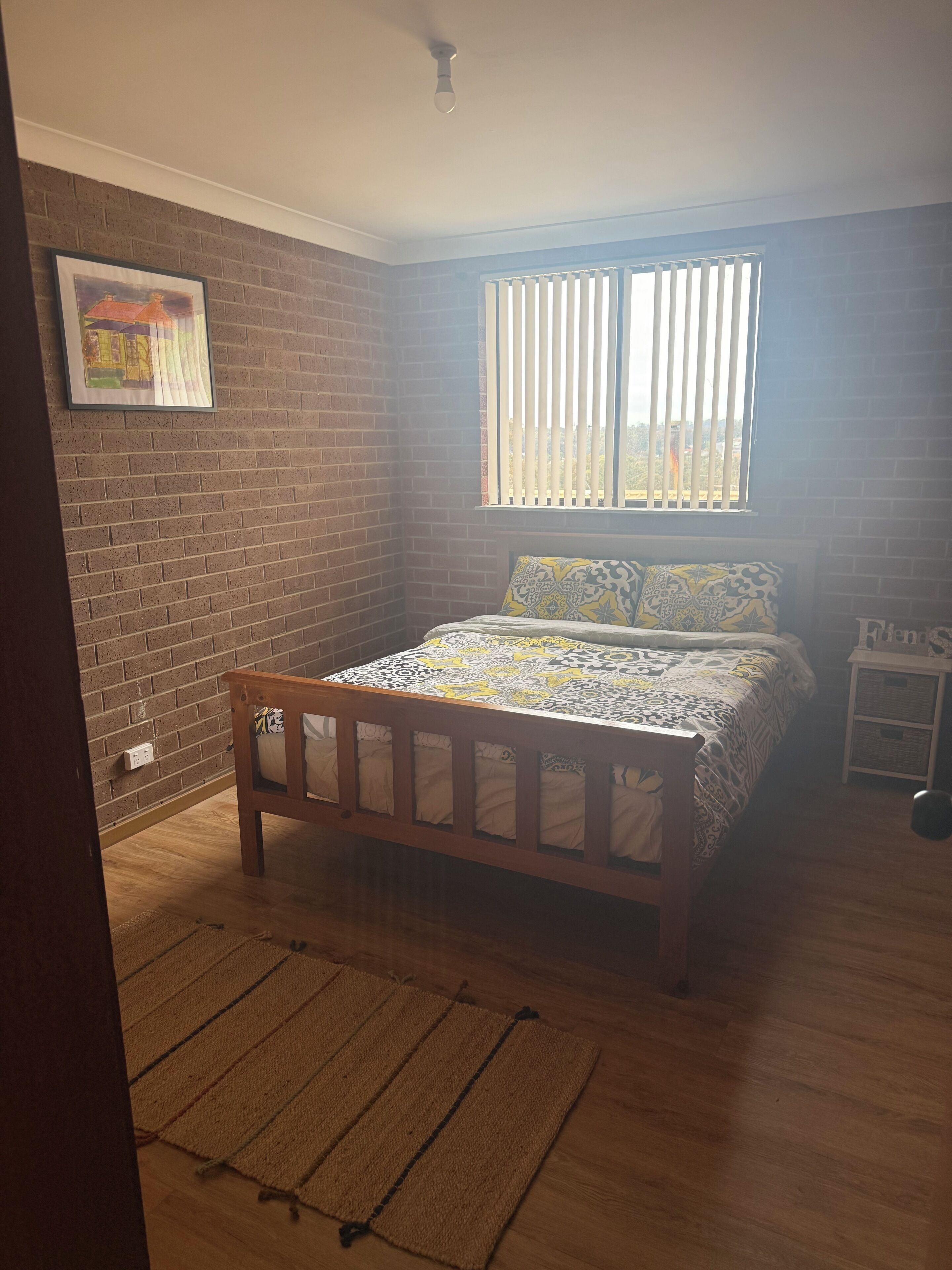Une chambre avec un lit en bois, un mur en briques, une fenêtre avec des stores et une petite table de chevet.