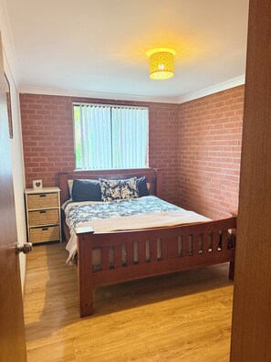 A bedroom with a wooden bed, a window with blinds, a bedside table with a basket, and a wall with a brick pattern. - The Lion’s Gate - family friendly 3-bedroom house moments from the beach (Catalina)