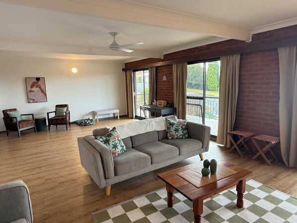 Un salon comprenant un canapé, des fauteuils, une table basse, un banc et une table sur laquelle se trouve une radio. Il y a une fenêtre avec des rideaux, un ventilateur de plafond et un tableau accroché au mur.