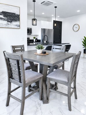 Een moderne eetruimte met een rechthoekige tafel en vier stoelen, een keuken op de achtergrond en een klok aan de muur.