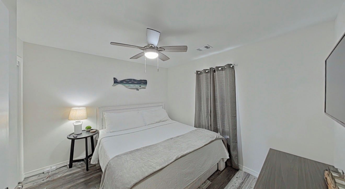Une chambre à coucher avec un lit, une table de chevet, une lampe, un ventilateur de plafond et une fenêtre avec des rideaux.