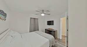 Ein Schlafzimmer mit Bett, Schreibtisch, Fernseher, Deckenventilator und einer Tür zu einem weiteren Raum.