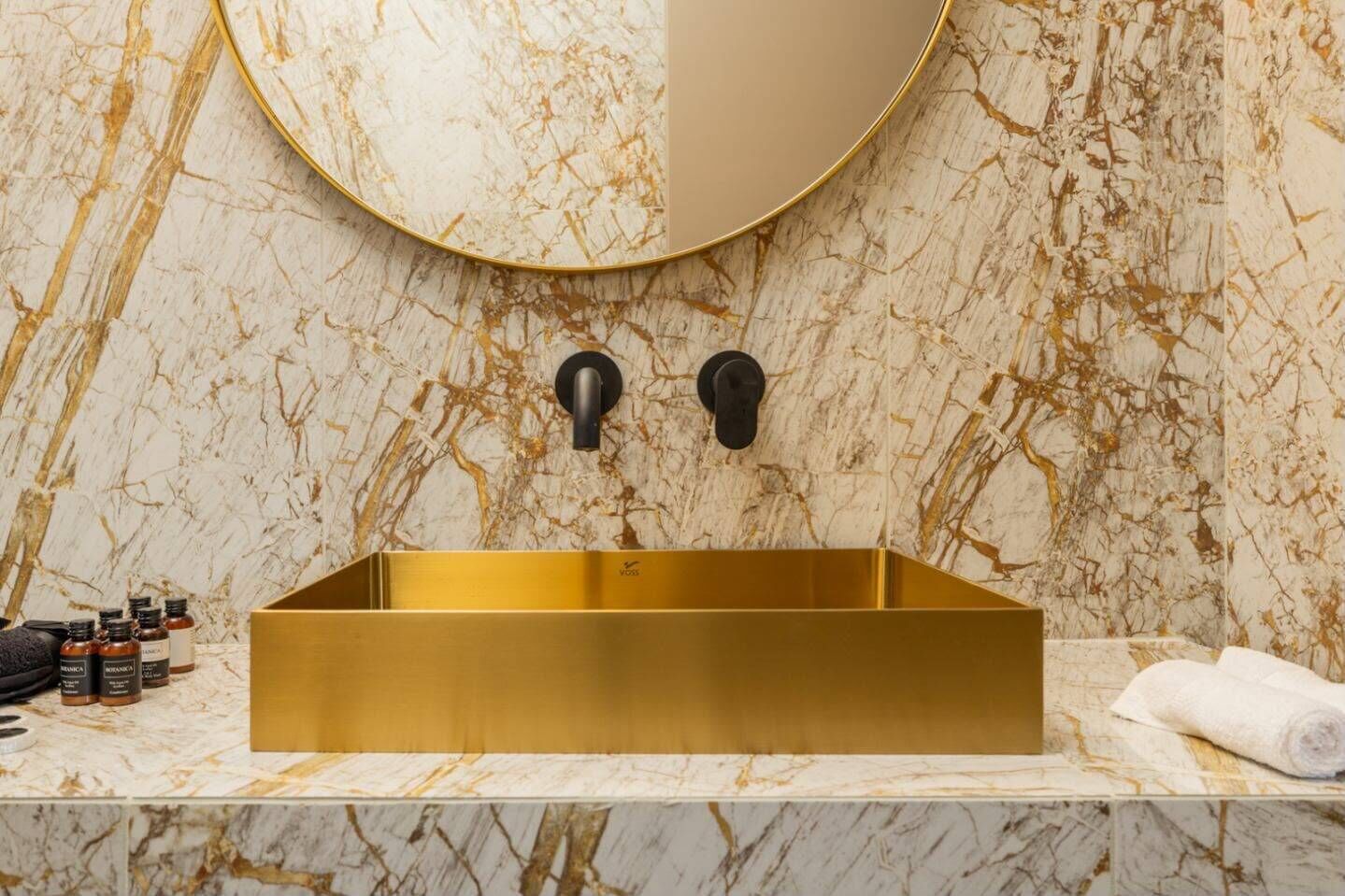 Ein Badezimmer mit einem rechteckigen, goldfarbenen Waschbecken, Marmorwänden und einem runden Spiegel.