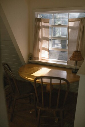 Quarto com mesa de madeira, duas cadeiras, janela com cortinas e abajur sobre a mesa.
