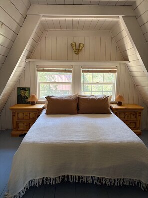 Ein Schlafzimmer mit schräg verlaufender Decke, zwei Fenstern mit Jalousien, einem Bett mit weißer Bettdecke, zwei Nachttischen mit Lampen und einer wandmontierten Leuchte.