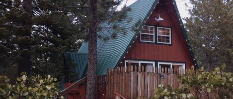 A cabin with a steeply pitched roof, wooden deck, and a fenced yard.