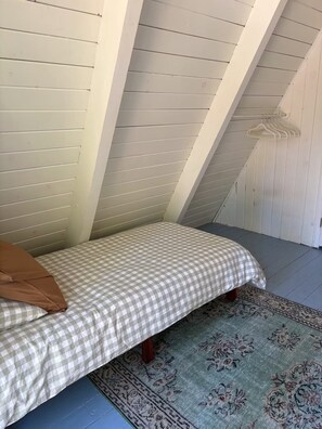 Quarto pequeno com uma cama de solteiro, colcha xadrez, tapete estampado e paredes de madeira branca.