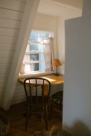 Ein Zimmer mit schrägen Wänden, einem Holztisch, einem Stuhl, einer Lampe und einem Fenster mit Vorhängen.