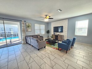 A living room with a sofa, chairs, a coffee table, a TV, and a ceiling fan. - 9BR ChampionsGate Villa | Private Pool & Spa | Game Room | Near Disney (Davenport)
