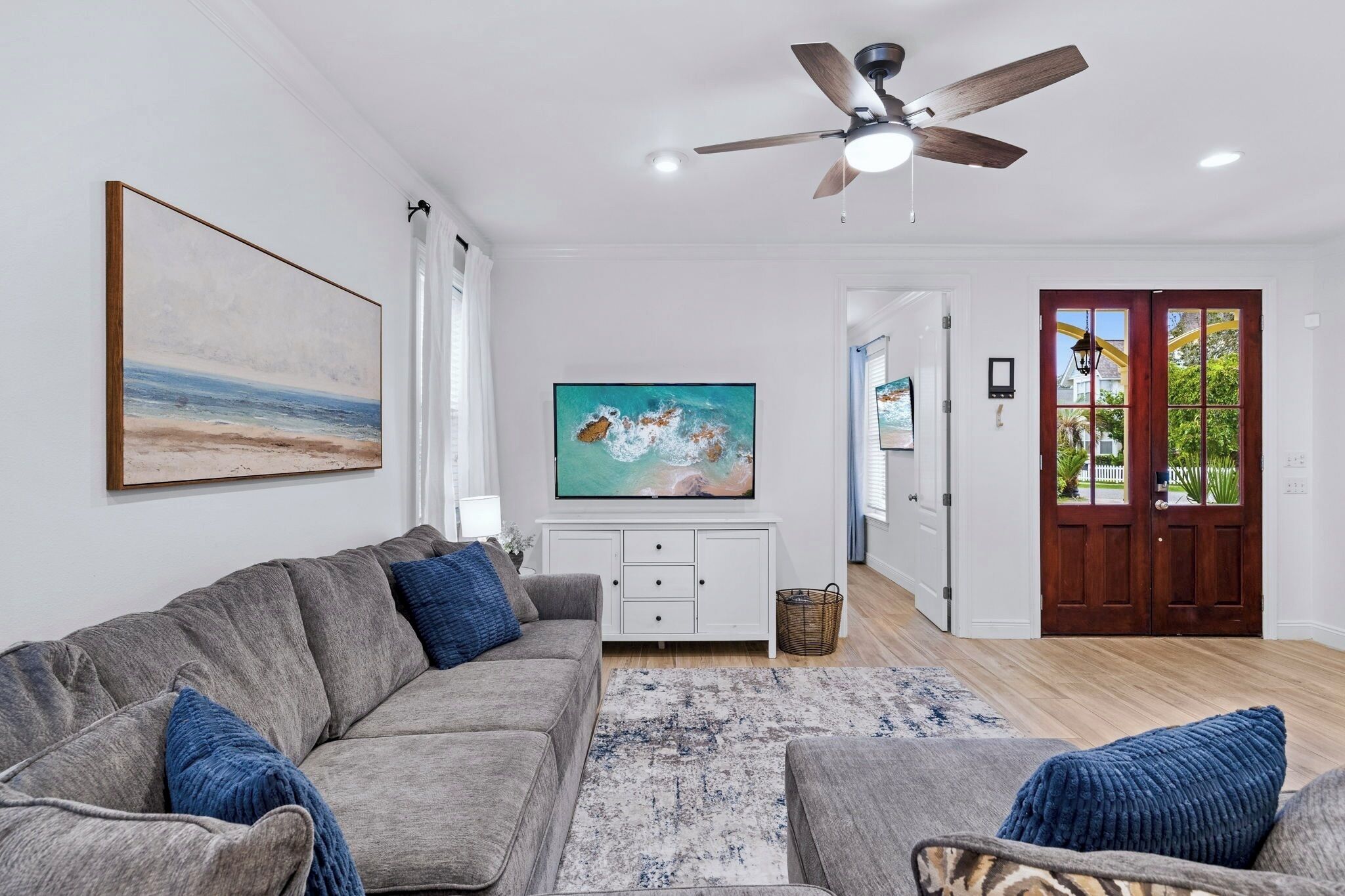A living room with a grey sofa, blue cushions, a flat-screen TV, a ceiling fan, and a wooden door leading outside.