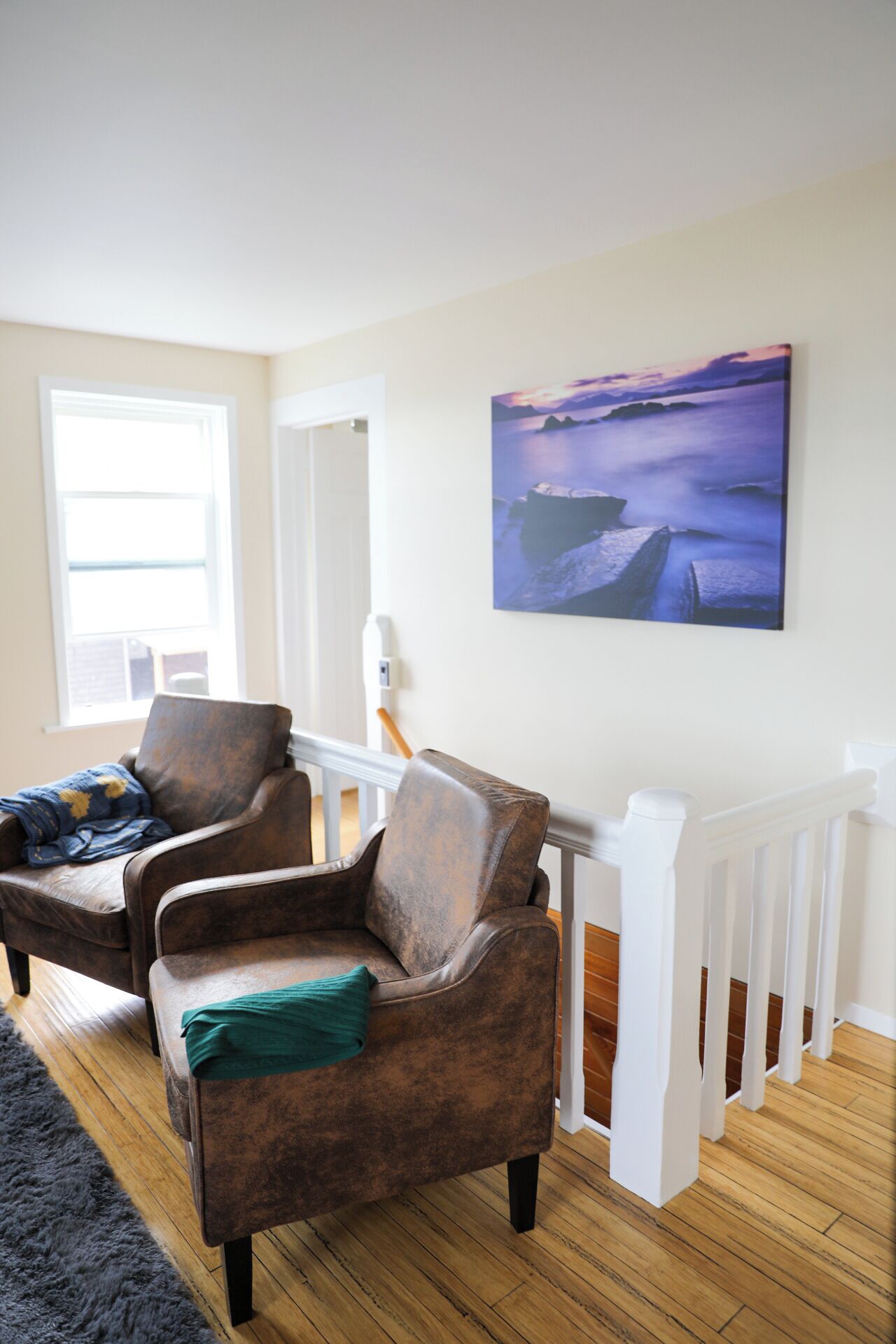 Uma sala de estar com duas poltronas de couro marrom, um corrimão branco e uma pintura de uma paisagem costeira.